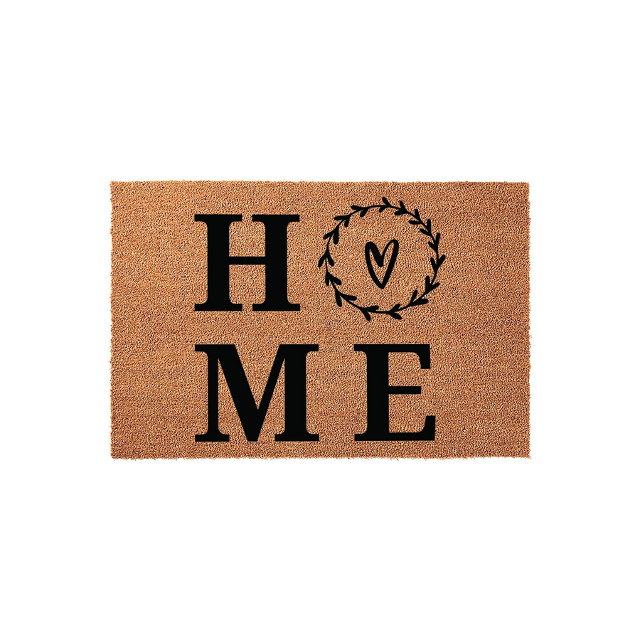 Home Coir Doormat featuring a rustic farmhouse design with the word HOME printed in bold black letters, where the letter O is replaced by a decorative leafy wreath surrounding a small hand-drawn heart on a natural tan fiber