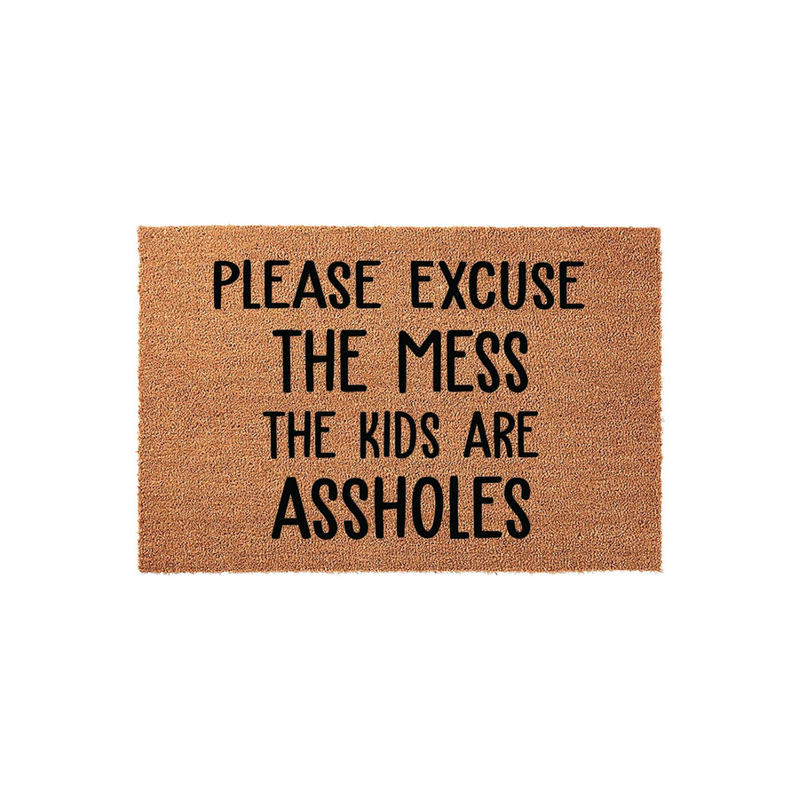 Funny coir doormat featuring the text Please Excuse The Mess The Kids Are Assholes in bold black lettering, set against a natural brown coconut fiber background for a humorous and welcoming entryway decor piece for any busy