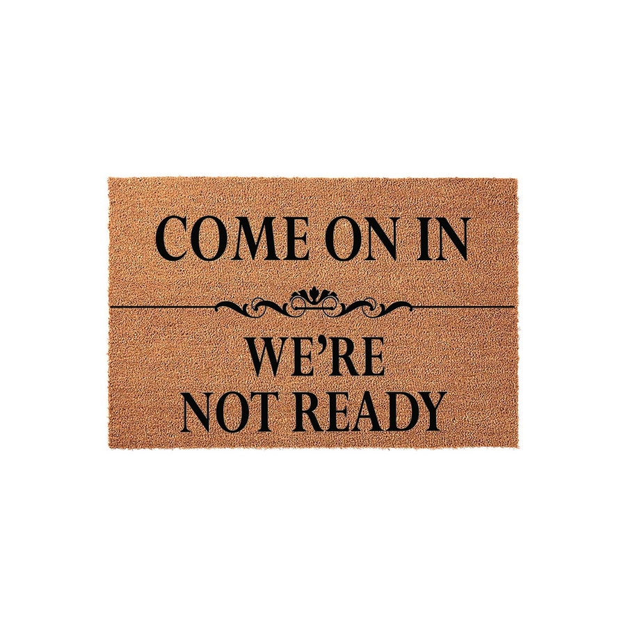Funny Come On In We Are Not Ready coir doormat featuring a natural brown fiber texture and bold black text with a decorative flourish, making it a humorous welcome mat for your front porch or entryway decor.