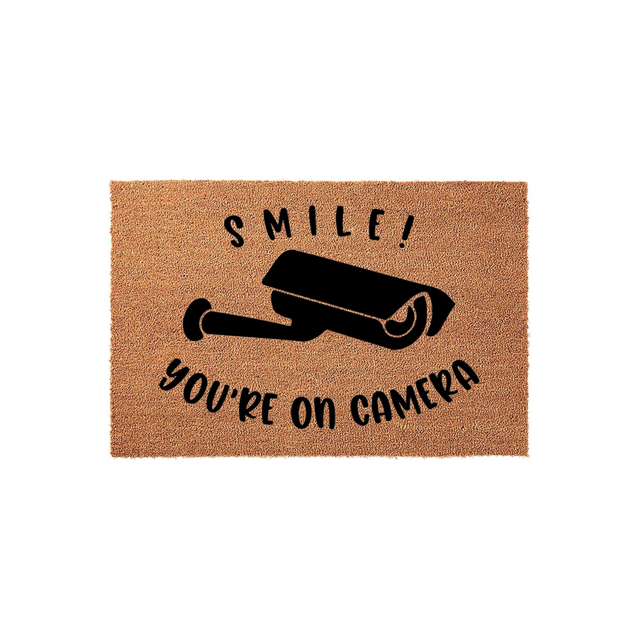 Smile You Are On Camera coir doormat featuring a black security camera graphic printed on a natural tan fiber background, creating a humorous and functional front porch welcome mat for your home entrance or security-conscious