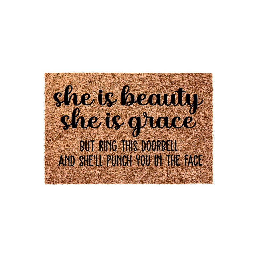 Funny doormat with a natural coir texture featuring the humorous quote she is beauty she is grace but ring this doorbell and she will punch you in the face printed in bold black lettering on a plain white studio background.