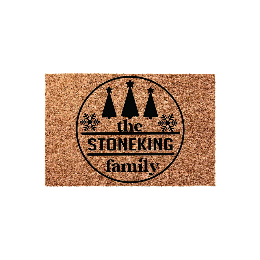 Personalized Christmas family name coir doormat featuring a black circular design with three pine trees, snowflakes, and custom text that reads the family name, set against a natural brown textured coir fiber background for