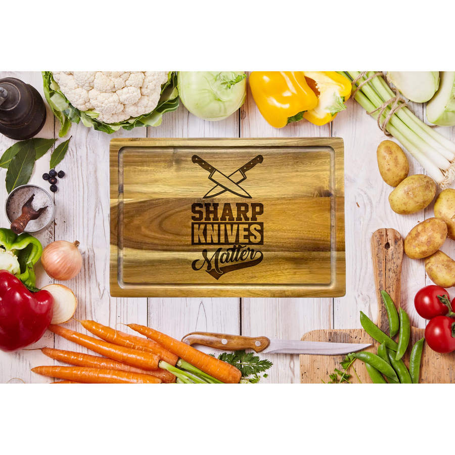 Engraved wooden cutting board featuring the text Sharp Knives Matter with crossed knife icons, centered on a rustic white wooden table surrounded by fresh vegetables like carrots, peppers, potatoes, and onions for a culinary