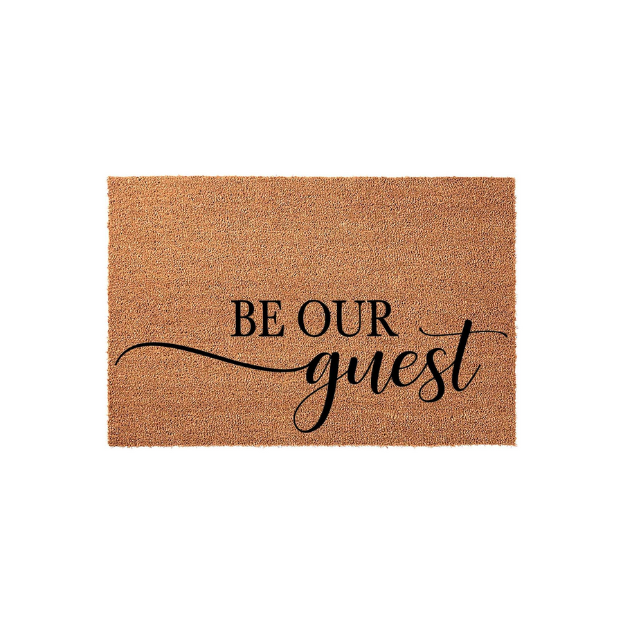 Be Our Guest coir doormat featuring a natural brown fiber texture and elegant black typography that reads Be Our Guest in a mix of bold block letters and a flowing script font, perfect for a welcoming front porch entryway decor.