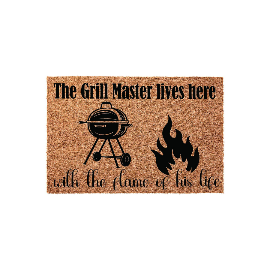 Grill Master coir doormat featuring a black graphic of a charcoal barbecue grill and a flame, with the text The Grill Master lives here with the flame of his life printed in a mix of bold and script fonts on a natural tan fiber