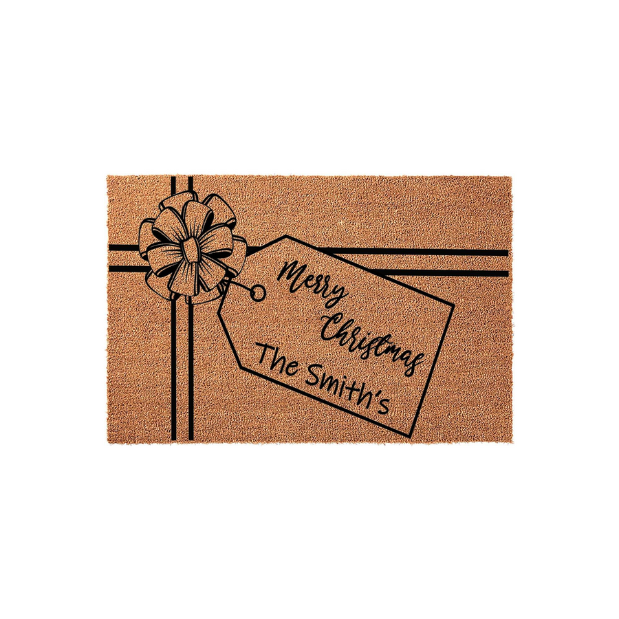 Personalized Merry Christmas doormat featuring a festive gift box design with a black bow and a hanging tag that says Merry Christmas and includes space for a family name on a natural coir fiber background for holiday home decor.