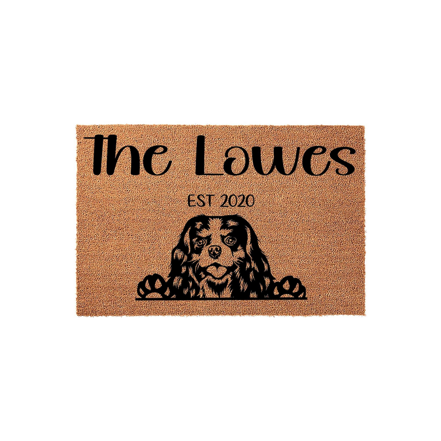 Personalized Cavalier King Charles Spaniel doormat featuring a custom family name and established year printed in black on a natural coir fiber mat, showcasing a cute dog peeking over the bottom edge with its paws visible.