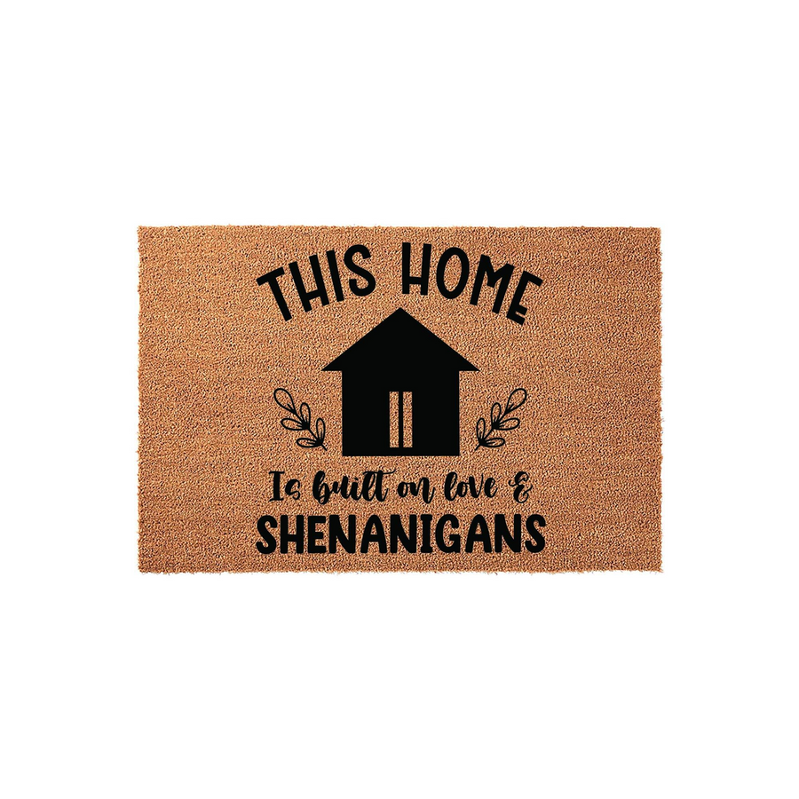 This Home Is Built On Love And Shenanigans coir doormat features a black house icon with leafy accents on a natural tan fiber background, creating a fun and welcoming entryway decor piece for any front porch or housewarming