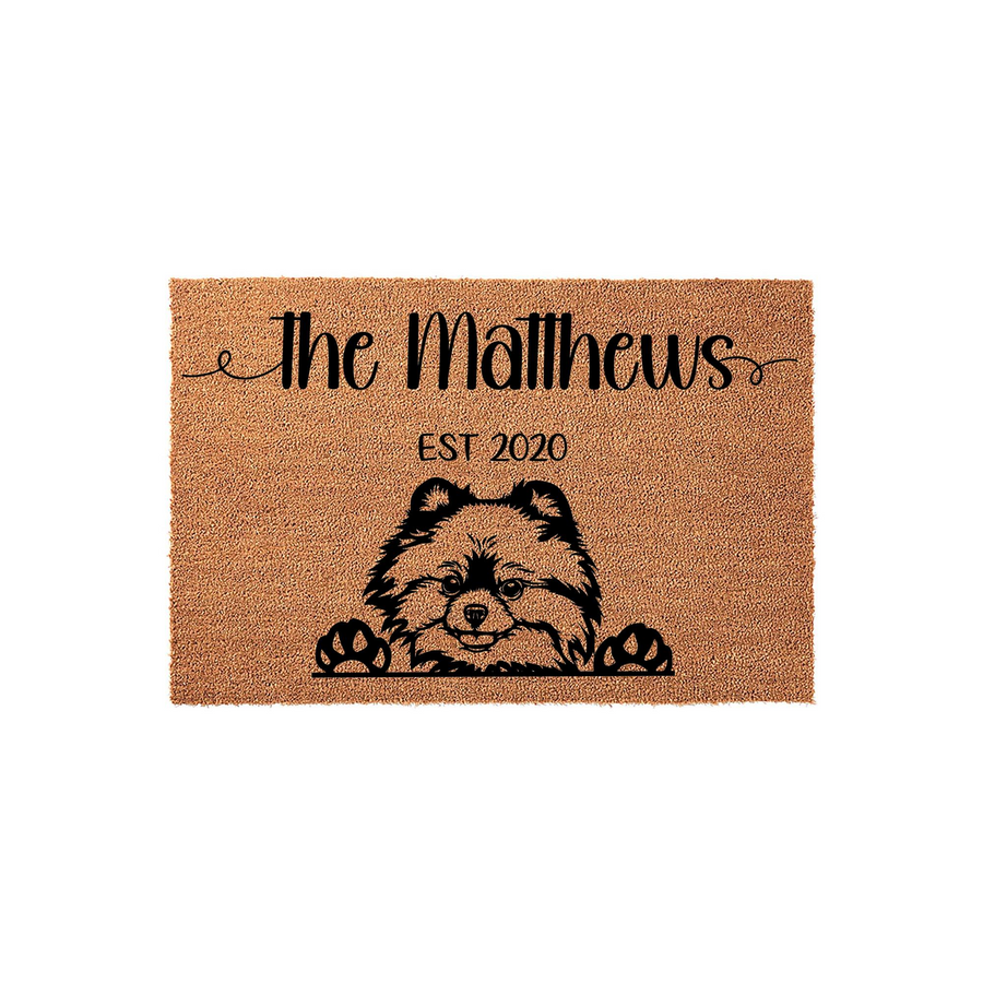 Personalized Pomeranian dog doormat featuring a cute illustrated dog peeking over the bottom edge, customized with a family name and established year on a natural tan coir fiber background for a welcoming front porch entrance.