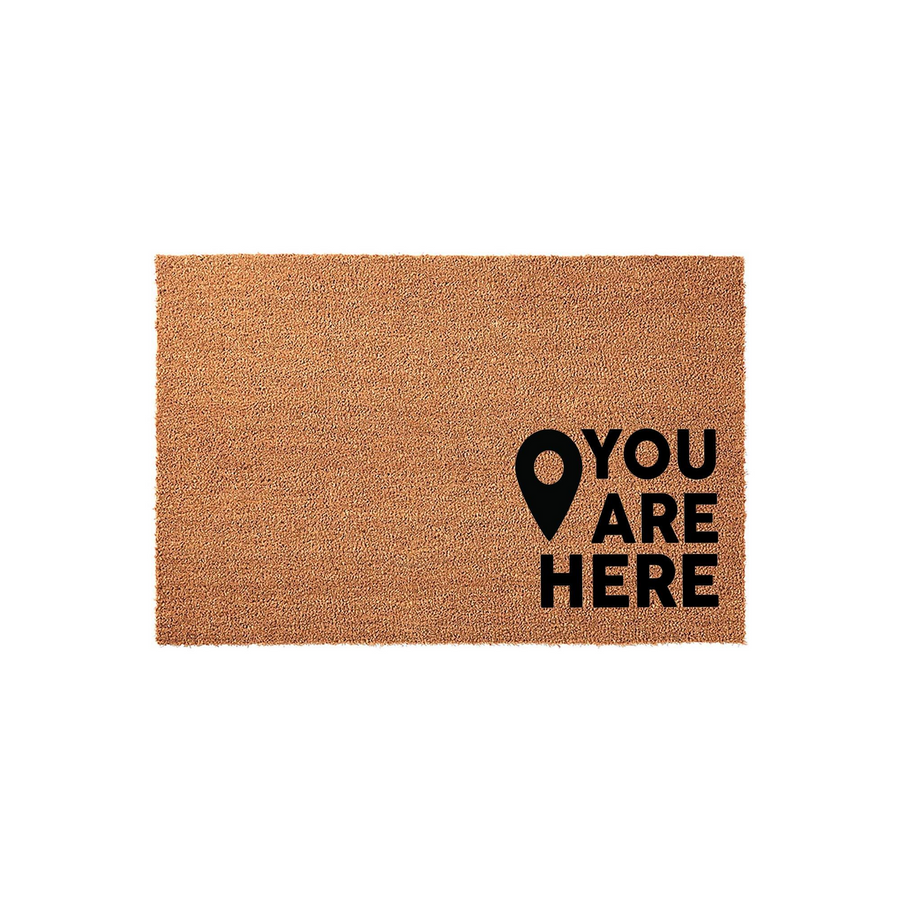 You Are Here coir doormat featuring a natural brown fiber texture and bold black text with a map location pin icon, providing a humorous and welcoming touch to your front porch entryway decor on a clean white studio background.