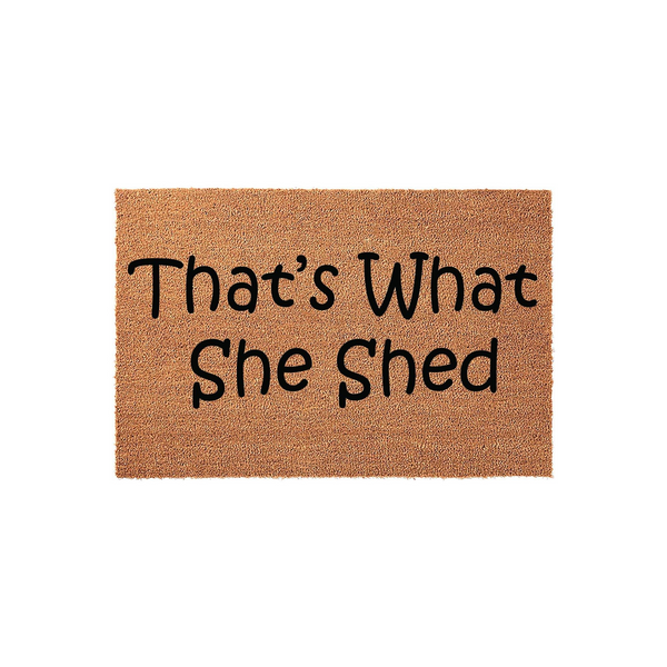 That's What She Shed funny doormat featuring a natural coir fiber texture and bold black lettering, designed as a humorous outdoor welcome mat for a backyard garden shed or a creative hobby space entrance on a plain white