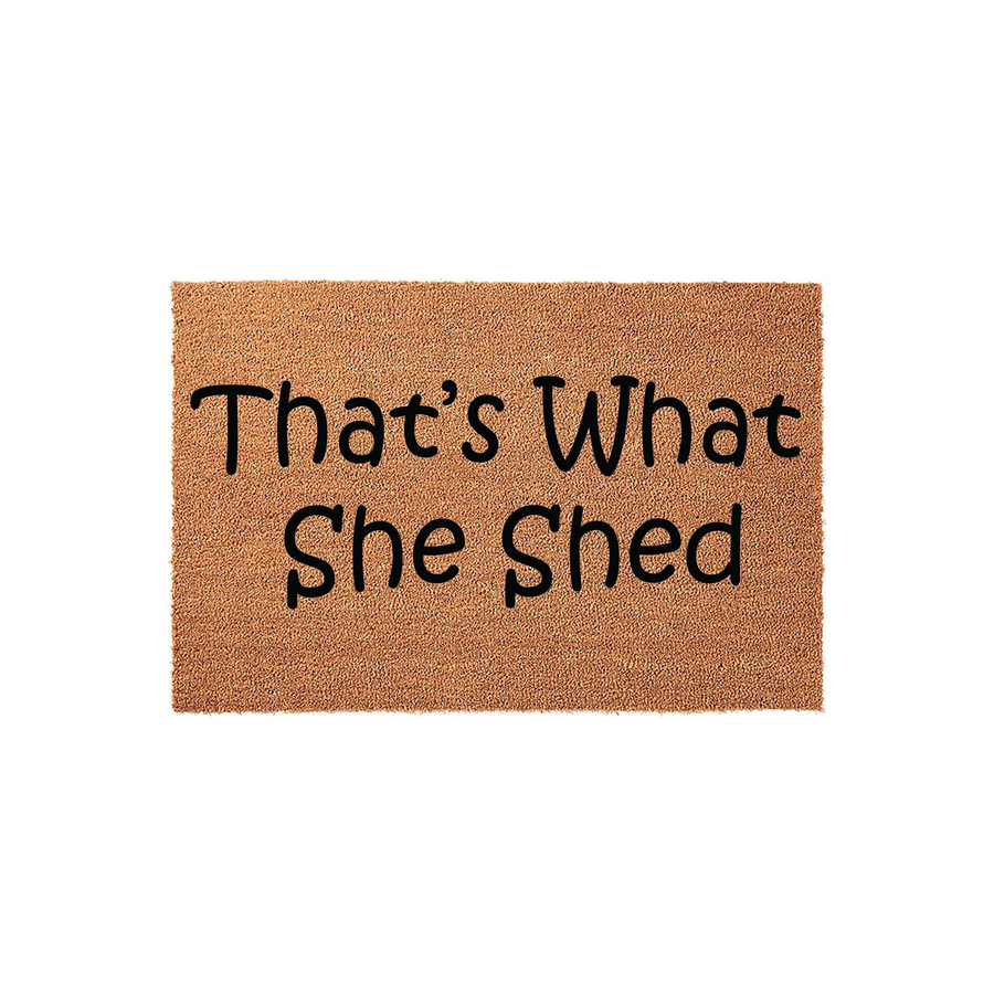That's What She Shed funny doormat featuring a natural coir fiber texture and bold black lettering, designed as a humorous outdoor welcome mat for a backyard garden shed or a creative hobby space entrance on a plain white