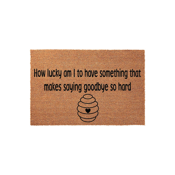 Natural coir doormat featuring the quote How lucky am I to have something that makes saying goodbye so hard printed in black text above a simple line-art beehive icon with a small heart in the center, set against a plain white