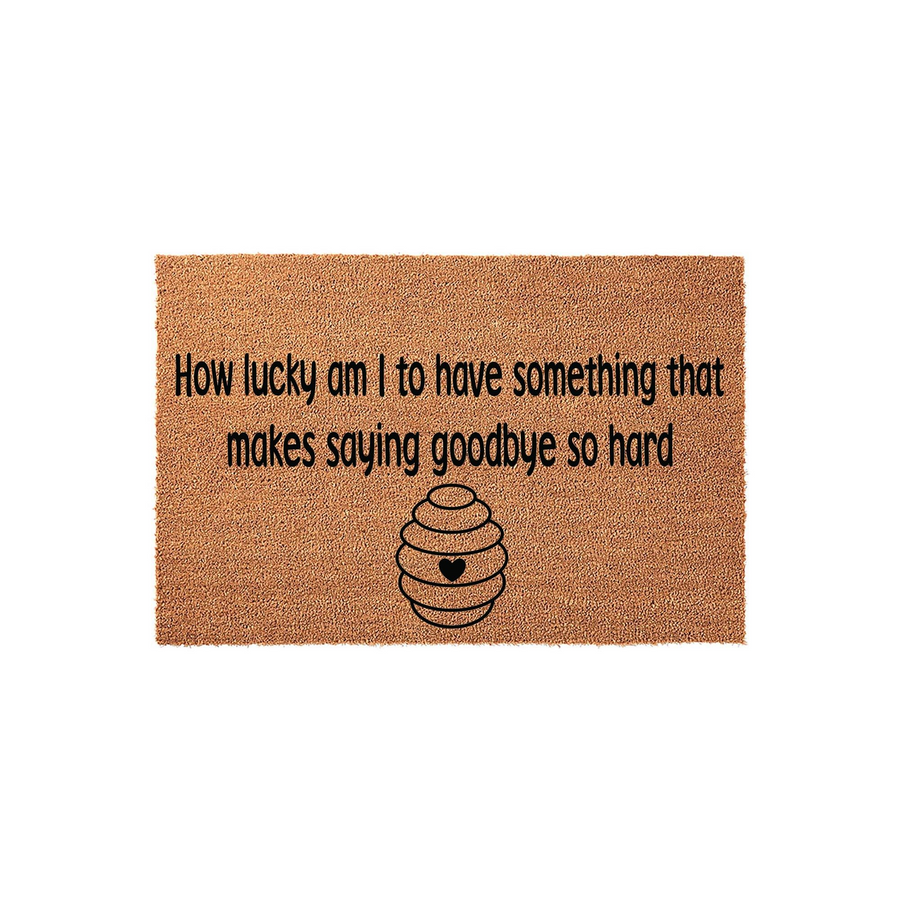 Natural coir doormat featuring the quote How lucky am I to have something that makes saying goodbye so hard printed in black text above a simple line-art beehive icon with a small heart in the center, set against a plain white