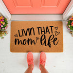Livin That Mom Life coir doormat for front porch entryways, featuring black script text and heart accents on a natural fiber welcome mat, shown on a white floor with pink sneakers and colorful flowers by a coral front door.