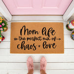 Mom Life doormat with the perfect mix of chaos and love text, a natural coir fiber welcome mat for front porch decor, featuring black script lettering on a tan rug placed on a white deck in front of a pink door for home entry.