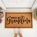World's Best Grandma coir doormat for front porch entry, featuring a natural fiber welcome mat with black script text and heart accents, placed on a white floor in front of a door with potted flowers and a woven basket nearby.