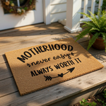 Motherhood doormat with a natural coir fiber texture, featuring bold black text that reads Motherhood never easy always worth it with a decorative arrow and heart design for a front porch welcome mat or home entryway decor.
