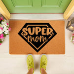 Super Mom doormat featuring a black superhero shield design on natural coir fiber, placed on a front porch with flowers and a lantern, making a fun welcome mat for a home entrance or a decorative outdoor floor rug for a mother.