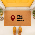 You Are Here doormat featuring a red map location pin icon on natural coir fiber, a funny front door welcome mat for home entryways that adds a humorous touch to your porch decor and makes a unique housewarming house sign.