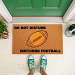 Football doormat with Do Not Disturb Watching Football text, a funny sports welcome mat for a front porch entrance, featuring a brown textured coir material with a graphic of a football for sports fans and home decor enthusiasts.