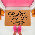 Funny Halloween doormat with bat shit crazy text and bat graphic, this natural coir welcome mat is a rude front porch decor piece for fall, featuring pumpkins and a bright pink door in the background for a festive seasonal look.