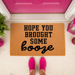 Funny welcome doormat with the text Hope You Brought Some Booze printed in black on a natural coir fiber mat, placed on a porch in front of a bright pink door with a person wearing pink sneakers standing on the mat for a home