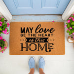 Coir welcome doormat featuring the quote May Love Be The Heart Of This Home in black text, placed on a porch with pink flowers and blue sneakers, making a stylish front door mat for housewarming or home decor entryways.