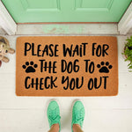 Funny dog welcome doormat featuring paw prints and text that reads Please Wait For The Dog To Check You Out, displayed on a front porch with a natural coir fiber texture and a light green door in the background for a pet lover