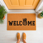 Pineapple welcome doormat featuring a natural coir fiber texture and bold black lettering, placed on a porch in front of a yellow door as a stylish outdoor floor mat for home entryways and front door decor for guests.