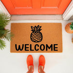 Pineapple welcome doormat for front porch, featuring a natural coir fiber texture with a black pineapple icon and welcome text, perfect for a housewarming entry rug or outdoor floor mat decor in front of a bright orange door.