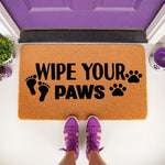 Wipe Your Paws doormat featuring black text and paw prints on a natural coir fiber mat, perfect for pet owners as a funny front porch welcome mat or entryway rug for dog lovers placed outside a purple front door with purple shoes.