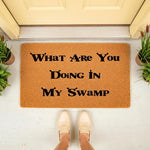 Funny doormat with the text What Are You Doing In My Swamp printed in black on a natural coir fiber welcome mat, placed on a porch in front of a door with a person wearing beige loafers standing on the mat for a humorous home