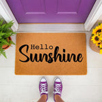 Hello Sunshine coir doormat featuring bold black script text on a natural fiber welcome mat, placed on a white porch in front of a purple door with sunflowers and a fern, creating a stylish entryway decor look for your home
