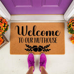 Funny Welcome To Our Nuthouse doormat with acorn design, a natural coir fiber welcome mat for front porch decor, featuring bold black text and fall foliage accents placed on a white floor in front of a purple door with orange