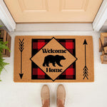 Bear Welcome Home doormat with red buffalo plaid pattern and black arrows, featuring a rustic cabin style outdoor welcome mat for front porch decor, placed on a white floor with wood accents and a person standing in beige shoes.