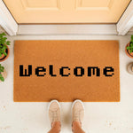 Pixel font Welcome doormat for front porch entry, featuring a natural coir fiber welcome mat with black blocky text, placed on a concrete doorstep with potted plants and a person wearing casual beige sneakers standing on the rug.