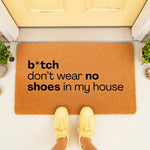 Funny doormat with rude text b*tch don't wear no shoes in my house, a natural coir welcome mat for front porch decor, featuring bold black lettering on a textured brown fiber rug surface for a humorous home entryway floor mat