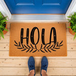 Hola welcome doormat with a natural coir fiber texture and black script lettering, featuring a decorative leaf design on a front porch with blue door and potted plants, perfect for a stylish entryway or housewarming home decor.