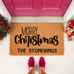 Personalized Merry Christmas doormat with family name, featuring a natural coir fiber welcome mat for holiday porch decor, placed in front of a bright pink door with festive pine greenery and wrapped gift boxes for a seasonal