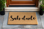 Sah dude coir doormat featuring bold black script text on a natural fiber welcome mat, placed on a gray tiled porch entry way with potted plants, perfect for a funny front door decor look or a casual housewarming entrance rug.