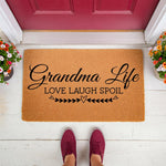 Grandma Life doormat for front porch, a natural coir welcome mat featuring black script text and heart accents, placed on a white wood floor by a red door with colorful potted flowers, perfect for a home entryway or housewarming