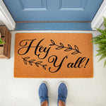 Hey Y'all coir doormat with black script text and leaf accents, a natural fiber welcome mat for front porch decor that adds a farmhouse touch to your entryway, shown on a porch with blue shoes and a blue door in the background.