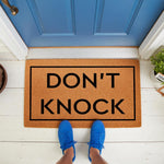 Don't Knock funny doormat featuring bold black text on a natural coir fiber welcome mat, placed on a white porch in front of a blue door with a person wearing blue sneakers standing on the mat for a humorous home entryway decor