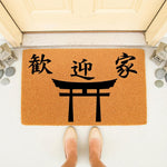 Asian style torii gate welcome doormat featuring black calligraphy on a natural coir fiber mat, placed at a front door entrance with decorative stone lanterns and smooth river rocks for a zen home decor look on a porch.