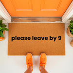Funny please leave by 9 doormat featuring bold black text on a natural coir fiber welcome mat, placed at an orange front door entrance with potted plants, perfect for a humorous home decor entryway or a quirky housewarming front