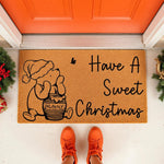 Christmas bear honey pot doormat featuring a cute cartoon bear in a Santa hat with a honey jar, perfect for a festive holiday welcome mat or front porch decor, shown on a doorstep with evergreen accents and bright orange sneakers.