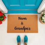 Nana and Grandpa's personalized coir doormat for front porch entry, featuring a natural fiber welcome mat design with black script text, placed on a white floor in front of a bright blue door with potted flowers on each side.