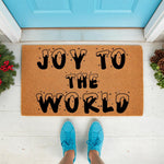 Joy to the World Christmas doormat made of natural coir fiber, featuring festive snowy lettering on a brown welcome mat placed on a porch with holiday greenery and pinecones for a seasonal front door entrance decor look.