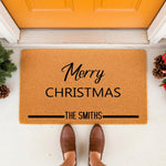 Personalized Merry Christmas doormat with family name, featuring a natural coir fiber welcome mat for front porch holiday decor, placed on a concrete entryway floor with festive pine cones and evergreen accents for a seasonal
